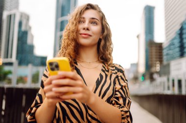 Telefonla gülümseyen genç bir kadının portresi. Metropolitan bir şehrin arka planında duruyor. Kıvırcık saçlı kadın blogları ya da açık hava metinleri. Yürümenin ve blog yazmanın kavramsal görüntüsü.