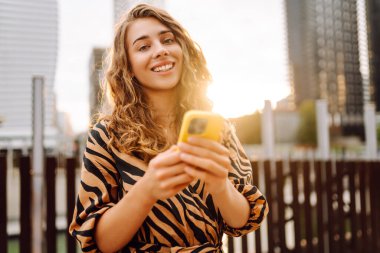 Telefonla gülümseyen genç bir kadının portresi. Metropolitan bir şehrin arka planında duruyor. Kıvırcık saçlı kadın blogları ya da açık hava metinleri. Yürümenin ve blog yazmanın kavramsal görüntüsü.