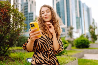 Telefonla gülümseyen genç bir kadının portresi. Metropolitan bir şehrin arka planında duruyor. Kıvırcık saçlı kadın blogları ya da açık hava metinleri. Yürümenin ve blog yazmanın kavramsal görüntüsü.