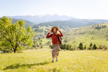 Şapka takan ve sırt çantası taşıyan kadın yürüyüşçü gün batımında yeşil tepede duruyor. Güzel bir kadın dağ manzarasını seviyor, yürüyüş parkurunda yürüyor. Seyahat kavramları, eğlence. Etkin yaşam biçimi.