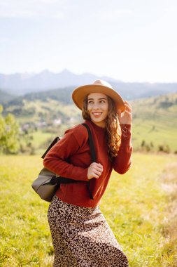 Şapka takan ve sırt çantası taşıyan kadın yürüyüşçü gün batımında yeşil tepede duruyor. Güzel bir kadın dağ manzarasını seviyor, yürüyüş parkurunda yürüyor. Seyahat kavramları, eğlence. Etkin yaşam biçimi.