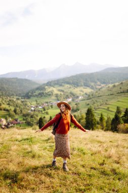 Şapka takan ve sırt çantası taşıyan kadın yürüyüşçü gün batımında yeşil tepede duruyor. Güzel bir kadın dağ manzarasını seviyor, yürüyüş parkurunda yürüyor. Seyahat kavramları, eğlence. Etkin yaşam biçimi.