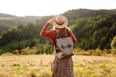 Şapka takan ve sırt çantası taşıyan kadın yürüyüşçü gün batımında yeşil tepede duruyor. Güzel bir kadın dağ manzarasını seviyor, yürüyüş parkurunda yürüyor. Seyahat kavramları, eğlence. Etkin yaşam biçimi.
