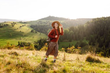 Şapka takan ve sırt çantası taşıyan kadın yürüyüşçü gün batımında yeşil tepede duruyor. Güzel bir kadın dağ manzarasını seviyor, yürüyüş parkurunda yürüyor. Seyahat kavramları, eğlence. Etkin yaşam biçimi.