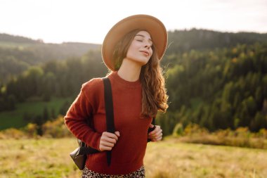 Şapka takan ve sırt çantası taşıyan kadın yürüyüşçü gün batımında yeşil tepede duruyor. Güzel bir kadın dağ manzarasını seviyor, yürüyüş parkurunda yürüyor. Seyahat kavramları, eğlence. Etkin yaşam biçimi.