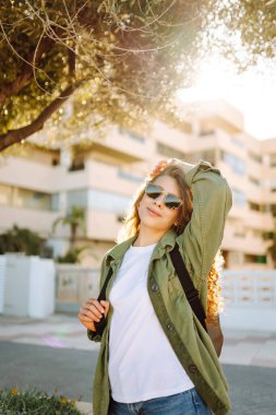 Neşeli bir kadın güneşli bir parkta eğleniyor. Güneş gözlüğü takan bir sırt çantası palmiye ağaçları arasında yürümekten zevk alır. Rahatlama, hafta sonları ve keyif kavramları.