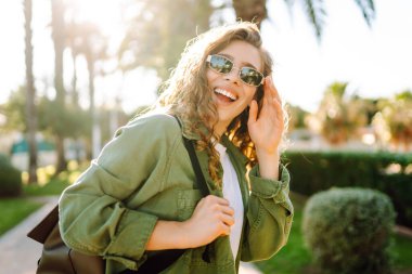 Neşeli bir kadın güneşli bir parkta eğleniyor. Güneş gözlüğü takan bir sırt çantası palmiye ağaçları arasında yürümekten zevk alır. Rahatlama, hafta sonları ve keyif kavramları.