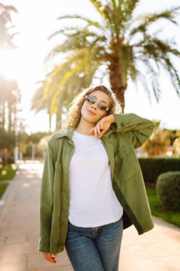 Neşeli bir kadın güneşli bir parkta eğleniyor. Güneş gözlüğü takan bir sırt çantası palmiye ağaçları arasında yürümekten zevk alır. Rahatlama, hafta sonları ve keyif kavramları.