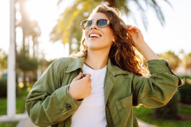 Neşeli bir kadın güneşli bir parkta eğleniyor. Güneş gözlüğü takan bir sırt çantası palmiye ağaçları arasında yürümekten zevk alır. Rahatlama, hafta sonları ve keyif kavramları.