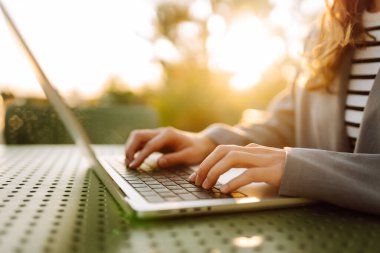 Gün batımında çatı terasında dizüstü bilgisayarda yazı yazan ellerin yakın çekimi. Diz üstü bilgisayarı olan serbest çalışan biri dışarıda çalışıyor. Uzaktan çalışma ve eğitim kavramı.