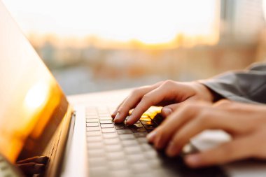 Gün batımında çatı terasında dizüstü bilgisayarda yazı yazan ellerin yakın çekimi. Diz üstü bilgisayarı olan serbest çalışan biri dışarıda çalışıyor. Uzaktan çalışma ve eğitim kavramı.