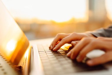 Gün batımında çatı terasında dizüstü bilgisayarda yazı yazan ellerin yakın çekimi. Diz üstü bilgisayarı olan serbest çalışan biri dışarıda çalışıyor. Uzaktan çalışma ve eğitim kavramı.