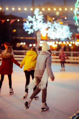 Genç bir kadın, parlak tatil ışıklarıyla süslenmiş bir kasaba meydanının arka planında paten kayıyor. Güzel bir kadın karlı bir günde paten kaymaktan hoşlanır. Konsept: Eğlence, mevsimlik.