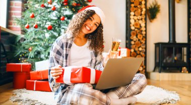 Bilgisayarlı ve yeni yıl hediyeli neşeli kadın internetten alışveriş yapıyor. Güzel bir kadın arka planda Noel ağacı olan süslü bir odada eğleniyor. Çevrimiçi alışveriş kavramı.