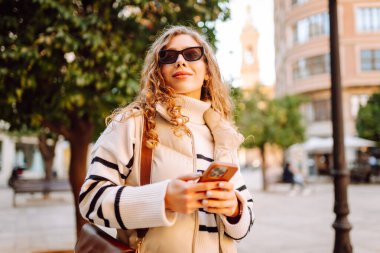 A portrait of a young woman standing in a town square with her phone at sunset. A beautiful woman enjoys a walk with her phone, texting, or blogging. Concepts: tourism, blogging. Active lifestyle.