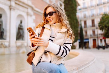 Gün batımında telefonuyla kasaba meydanında duran genç bir kadının portresi. Güzel bir kadın telefonuyla, mesajlarıyla ya da bloguyla yürümekten hoşlanır. Konseptler: Turizm, blog. Etkin yaşam biçimi.