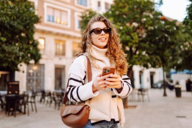 Gün batımında telefonuyla kasaba meydanında duran genç bir kadının portresi. Güzel bir kadın telefonuyla, mesajlarıyla ya da bloguyla yürümekten hoşlanır. Konseptler: Turizm, blog. Etkin yaşam biçimi.