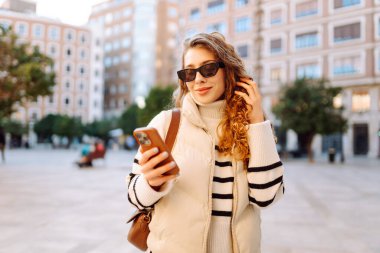 Gün batımında telefonuyla kasaba meydanında duran genç bir kadının portresi. Güzel bir kadın telefonuyla, mesajlarıyla ya da bloguyla yürümekten hoşlanır. Konseptler: Turizm, blog. Etkin yaşam biçimi.