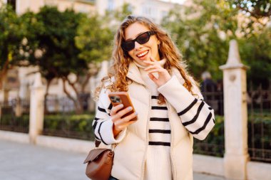 Gün batımında telefonuyla kasaba meydanında duran genç bir kadının portresi. Güzel bir kadın telefonuyla, mesajlarıyla ya da bloguyla yürümekten hoşlanır. Konseptler: Turizm, blog. Etkin yaşam biçimi.