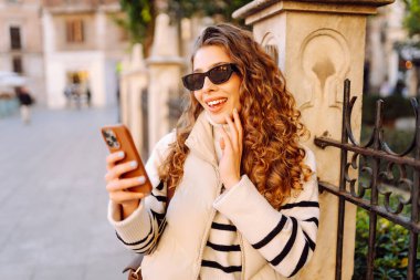 Gün batımında telefonuyla kasaba meydanında duran genç bir kadının portresi. Güzel bir kadın telefonuyla, mesajlarıyla ya da bloguyla yürümekten hoşlanır. Konseptler: Turizm, blog. Etkin yaşam biçimi.