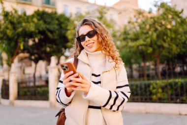 Gün batımında telefonuyla kasaba meydanında duran genç bir kadının portresi. Güzel bir kadın telefonuyla, mesajlarıyla ya da bloguyla yürümekten hoşlanır. Konseptler: Turizm, blog. Etkin yaşam biçimi.