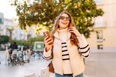 Gün batımında telefonuyla kasaba meydanında duran genç bir kadının portresi. Güzel bir kadın telefonuyla, mesajlarıyla ya da bloguyla yürümekten hoşlanır. Konseptler: Turizm, blog. Etkin yaşam biçimi.