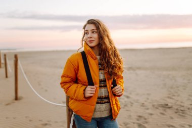 A young traveler in a bright jacket and with a backpack strolls along a sandy beach at sunset. The happy woman enjoys the soft sun by the water. Concept: nature, adventure.
