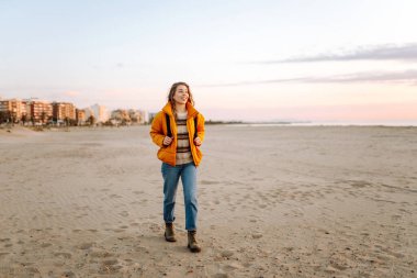 Parlak ceketli genç bir gezgin ve gün batımında kumsalda sırt çantasıyla geziniyor. Mutlu kadın suyun kenarındaki yumuşak güneşin tadını çıkarıyor. Kavram: doğa, macera.