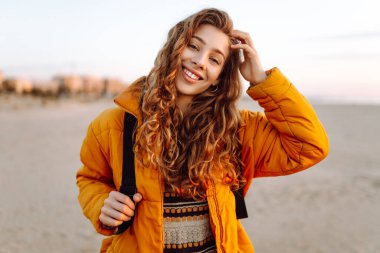 A young traveler in a bright jacket and with a backpack strolls along a sandy beach at sunset. The happy woman enjoys the soft sun by the water. Concept: nature, adventure.