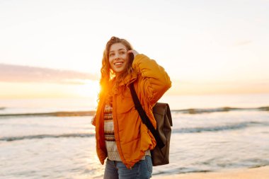 Parlak ceketli genç bir gezgin ve gün batımında kumsalda sırt çantasıyla geziniyor. Mutlu kadın suyun kenarındaki yumuşak güneşin tadını çıkarıyor. Kavram: doğa, macera.