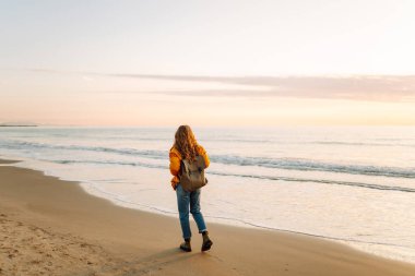 Parlak ceketli genç bir gezgin ve gün batımında kumsalda sırt çantasıyla geziniyor. Mutlu kadın suyun kenarındaki yumuşak güneşin tadını çıkarıyor. Kavram: doğa, macera.