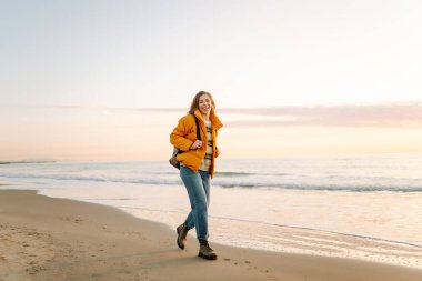 Parlak ceketli genç bir gezgin ve gün batımında kumsalda sırt çantasıyla geziniyor. Mutlu kadın suyun kenarındaki yumuşak güneşin tadını çıkarıyor. Kavram: doğa, macera.