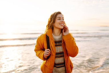 Parlak ceketli genç bir gezgin ve gün batımında kumsalda sırt çantasıyla geziniyor. Mutlu kadın suyun kenarındaki yumuşak güneşin tadını çıkarıyor. Kavram: doğa, macera.