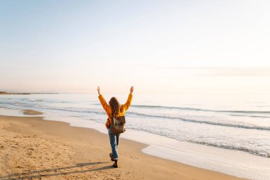 Parlak ceketli genç bir gezgin ve gün batımında kumsalda sırt çantasıyla geziniyor. Mutlu kadın suyun kenarındaki yumuşak güneşin tadını çıkarıyor. Kavram: doğa, macera.
