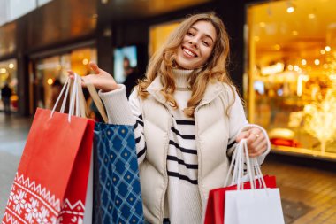 Parlak bir Noel sokağında elinde poşetlerle genç bir kadının portresi. Mutlu bir kadın parlak ışıkların ve Noel alışverişinin tadını çıkarır. Rahatlama, kutlama ve yaşam tarzı kavramı.