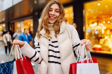 Parlak bir Noel sokağında elinde poşetlerle genç bir kadının portresi. Mutlu bir kadın parlak ışıkların ve Noel alışverişinin tadını çıkarır. Rahatlama, kutlama ve yaşam tarzı kavramı.