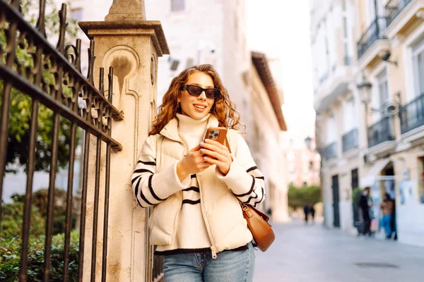 Gün batımında telefonuyla kasaba meydanında duran genç bir kadının portresi. Güzel bir kadın telefonuyla, mesajlarıyla ya da bloguyla yürümekten hoşlanır. Konseptler: Turizm, blog. Etkin yaşam biçimi.