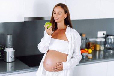 Modern bir mutfakta elinde elma tutan hamile bir kadın. Gülümseyen bir kadın güneşli bir günde sıcak bir odada yemek ya da atıştırmalık hazırlar. Sağlıklı beslenme, hamilelik kavramları. Yaşam biçimi.