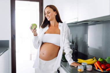 Modern bir mutfakta elinde elma tutan hamile bir kadın. Gülümseyen bir kadın güneşli bir günde sıcak bir odada yemek ya da atıştırmalık hazırlar. Sağlıklı beslenme, hamilelik kavramları. Yaşam biçimi.