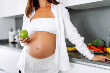 Modern bir mutfakta elinde elma tutan hamile bir kadın. Gülümseyen bir kadın güneşli bir günde sıcak bir odada yemek ya da atıştırmalık hazırlar. Sağlıklı beslenme, hamilelik kavramları. Yaşam biçimi.