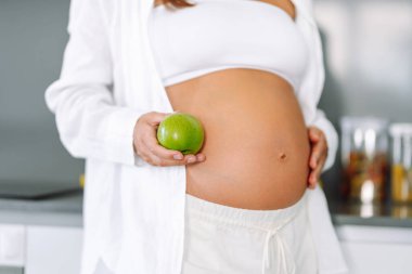 Modern bir mutfakta elinde elma tutan hamile bir kadın. Gülümseyen bir kadın güneşli bir günde sıcak bir odada yemek ya da atıştırmalık hazırlar. Sağlıklı beslenme, hamilelik kavramları. Yaşam biçimi.