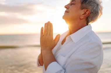 Gri saçlı bir kadın özgür hissediyor kumlu bir sahilde günbatımının tadını çıkarıyor. Olgun bir kadın güneş ışınlarıyla denizde gezinir. Mutlu bir emeklilik. Özgürlük kavramı, keyif. Yaşam biçimi.