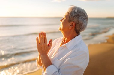 Gri saçlı bir kadın özgür hissediyor kumlu bir sahilde günbatımının tadını çıkarıyor. Olgun bir kadın güneş ışınlarıyla denizde gezinir. Mutlu bir emeklilik. Özgürlük kavramı, keyif. Yaşam biçimi.