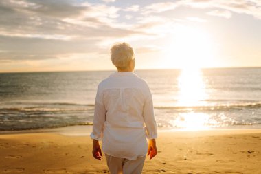 Gri saçlı bir kadın özgür hissediyor kumlu bir sahilde günbatımının tadını çıkarıyor. Olgun bir kadın güneş ışınlarıyla denizde gezinir. Mutlu bir emeklilik. Özgürlük kavramı, keyif. Yaşam biçimi.