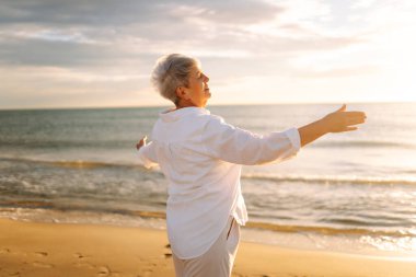 Gri saçlı bir kadın özgür hissediyor kumlu bir sahilde günbatımının tadını çıkarıyor. Olgun bir kadın güneş ışınlarıyla denizde gezinir. Mutlu bir emeklilik. Özgürlük kavramı, keyif. Yaşam biçimi.