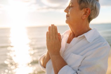 Gri saçlı bir kadın özgür hissediyor kumlu bir sahilde günbatımının tadını çıkarıyor. Olgun bir kadın güneş ışınlarıyla denizde gezinir. Mutlu bir emeklilik. Özgürlük kavramı, keyif. Yaşam biçimi.