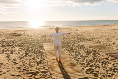 Gri saçlı bir kadın özgür hissediyor kumlu bir sahilde günbatımının tadını çıkarıyor. Olgun bir kadın güneş ışınlarıyla denizde gezinir. Mutlu bir emeklilik. Özgürlük kavramı, keyif. Yaşam biçimi.