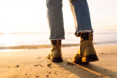 Güneş ışınlarında denizin arka planına karşı çizmeli bir kadının bacaklarının yakın çekimi. Günbatımında botlarıyla kumlu bir plajda duran bir insan. Yürüyüş, macera ve yürüyüş kavramları.