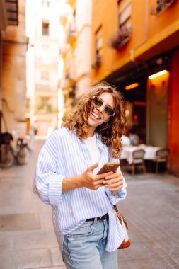 Telefonu olan neşeli bir kadın güneşli bir günde şehir sokaklarında geziniyor. Güzel bir turist güneşli bir günün ve telefonuyla blog yazmanın tadını çıkarır. Turizm, tatil ve hafta sonları için konseptler.