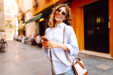 Telefonu olan neşeli bir kadın güneşli bir günde şehir sokaklarında geziniyor. Güzel bir turist güneşli bir günün ve telefonuyla blog yazmanın tadını çıkarır. Turizm, tatil ve hafta sonları için konseptler.